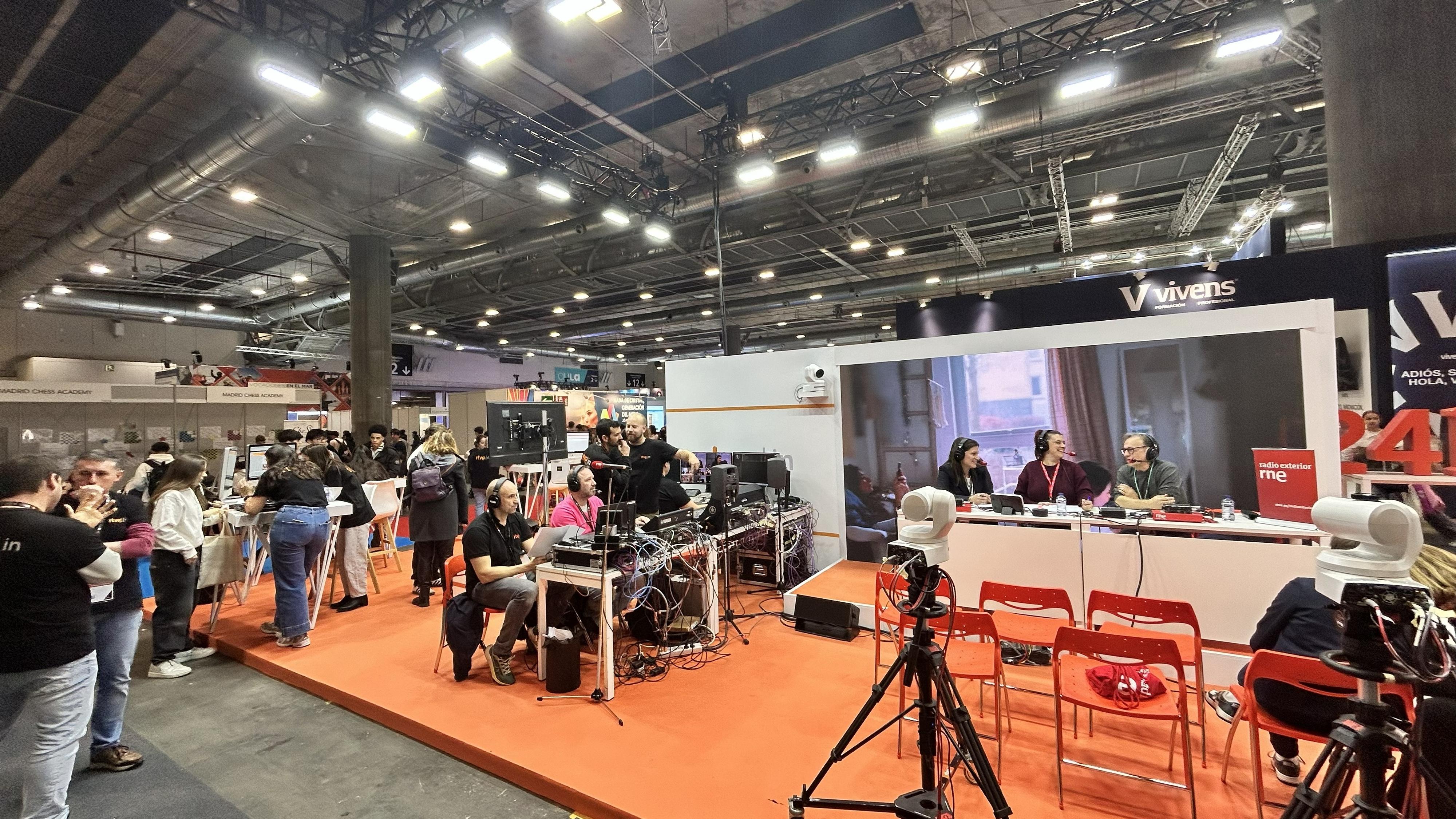 Stand de RTVEin en Aula 2026 En un recinto ferial, un set de grabación con cámaras y micrófonos es el centro de atención. Tres personas están sentadas a una mesa con el logo "radio marca me", posiblemente grabando un programa. Otros stands y personas interactuando completan la escena.