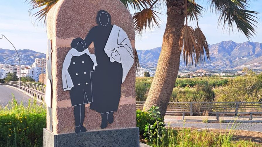 Monumento en Torre del Mar a las víctimas de la Desbandá. Siluetas recortadas de adulto y niño abrazados. Fondo con palmera, carretera y edificios.
