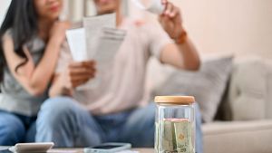 A close-up image of a jar of coins on a coffee table with a blurred background of a stressed couple worrying about their household bills and planning their finances. savings, budget, income