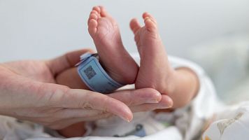 A baby's feet are held up by a person. The baby is wearing a blue tag. The baby is in a hospital bed