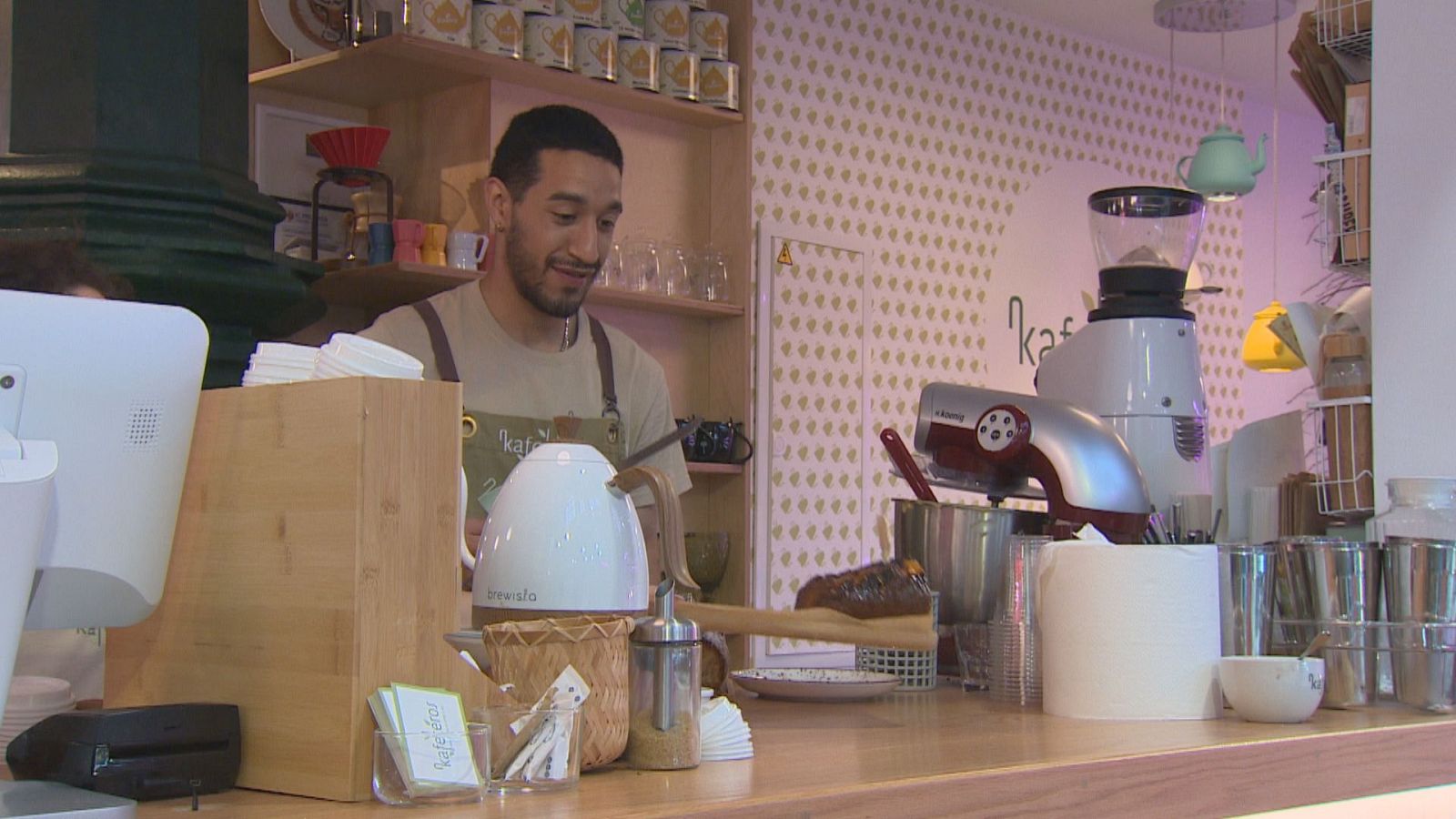 Barista sonriente con delantal "kafé" detrás de un mostrador lleno de equipo de cafetería, incluyendo una tetera "brewista" y un molinillo de café. Fondo con decoración dorada y estanterías.