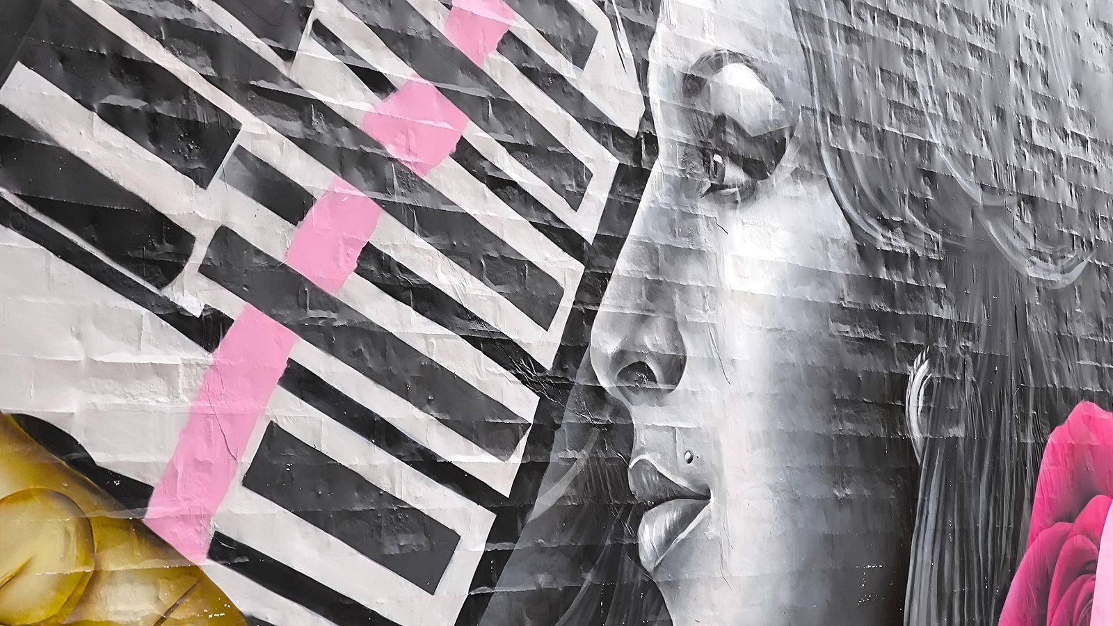 Mural de Amy Winehouse en pared de ladrillo con patrón geométrico, formas rosas y flor roja. Destaca el perfil de la cantante con piercing.