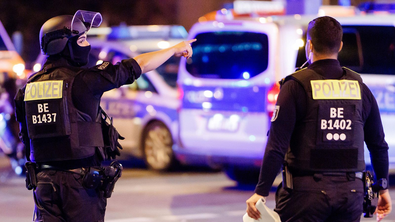 Dos policías alemanes, "POLIZEI" BE B1437 y "POLIZEI" BE B1402, en una calle nocturna. Uno señala, el otro sostiene un objeto blanco. Luces azules de emergencia al fondo.