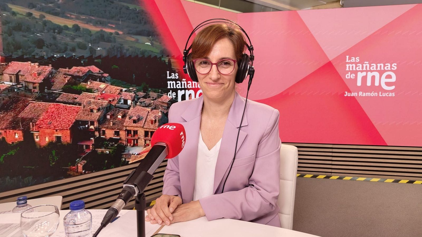 Pepa Fernández, periodista española, sonríe en un estudio de radio con gafas rojas, americana lila y auriculares. En la mesa, un micrófono "rne" y botellas. Fondo con pueblo rural y "Las mañanas de RNE".