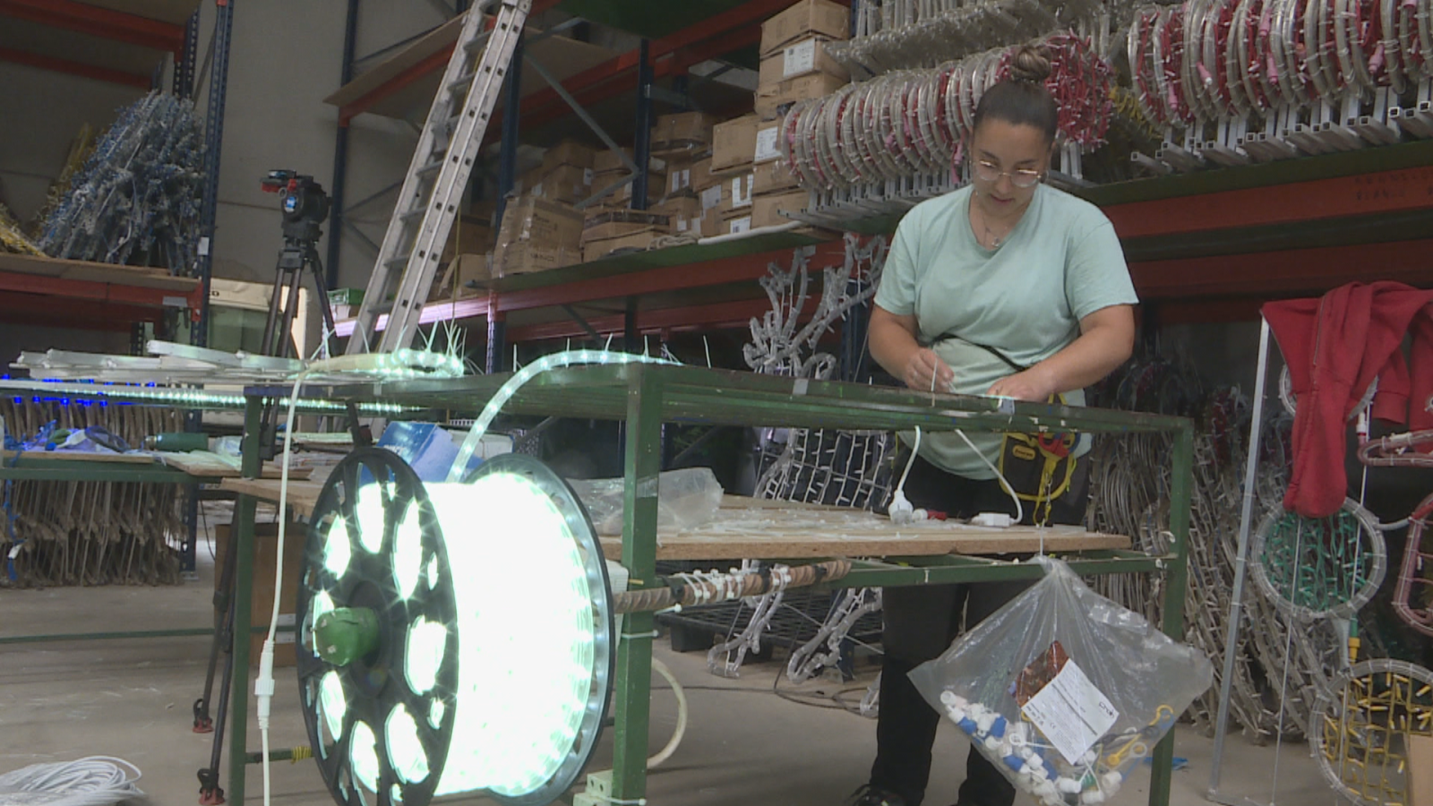 Mujer en taller industrial preparando luces LED blancas y azules. Estanterías con decoraciones navideñas al fondo. Cámara de vídeo grabando.