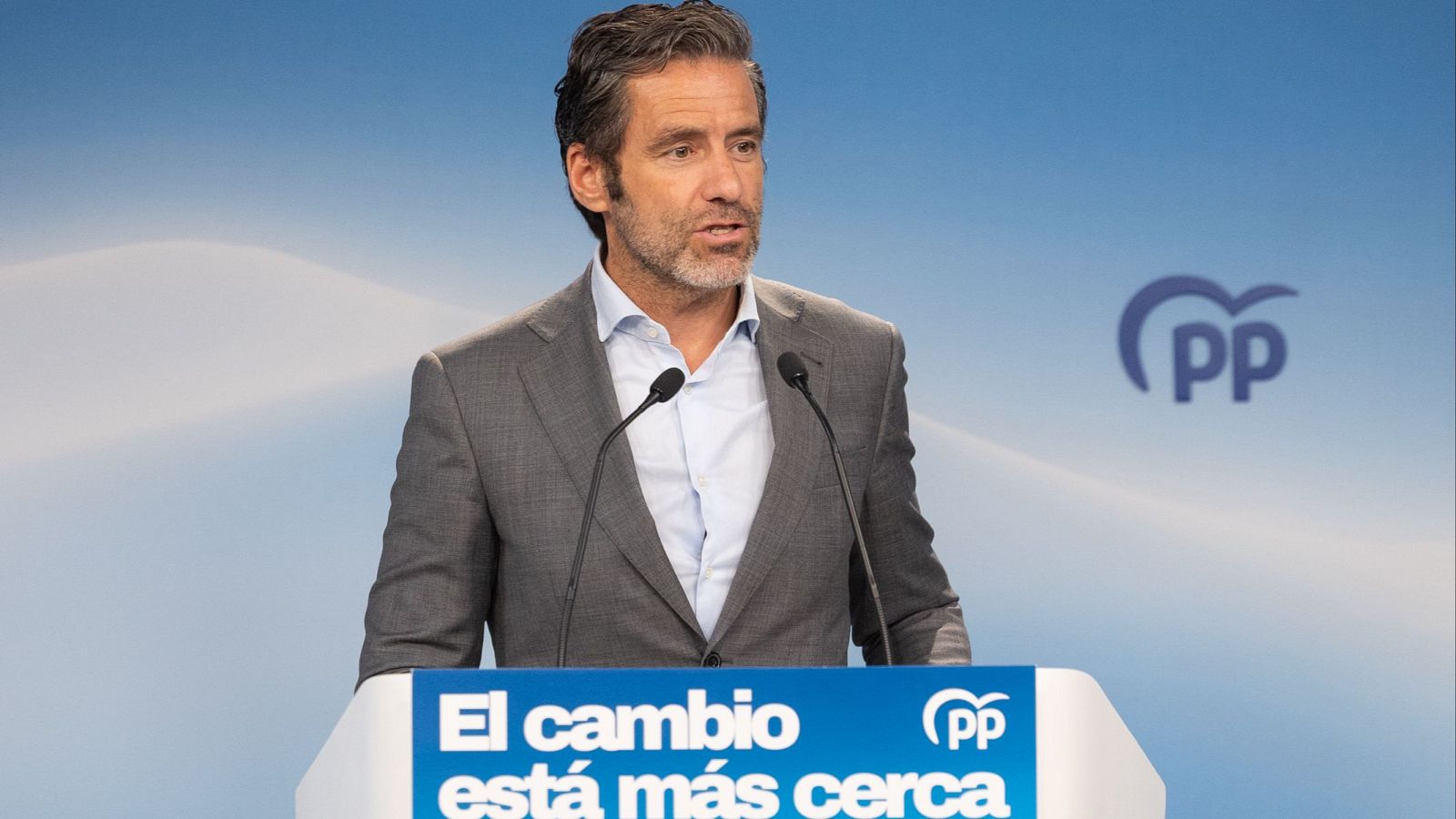 Borja Semper, portavoz del PP, habla en un atril con el eslogan "El cambio está más cerca" y el logo del partido. Viste traje gris y camisa azul. Fondo azul degradado con logo del PP.