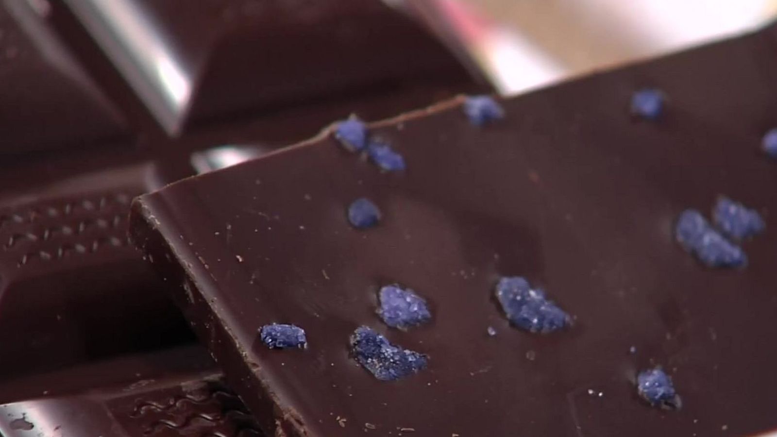 Chocolate oscuro con cristales azul violáceo esparcidos sobre su superficie lisa y brillante. Otros trozos de chocolate desenfocados al fondo.