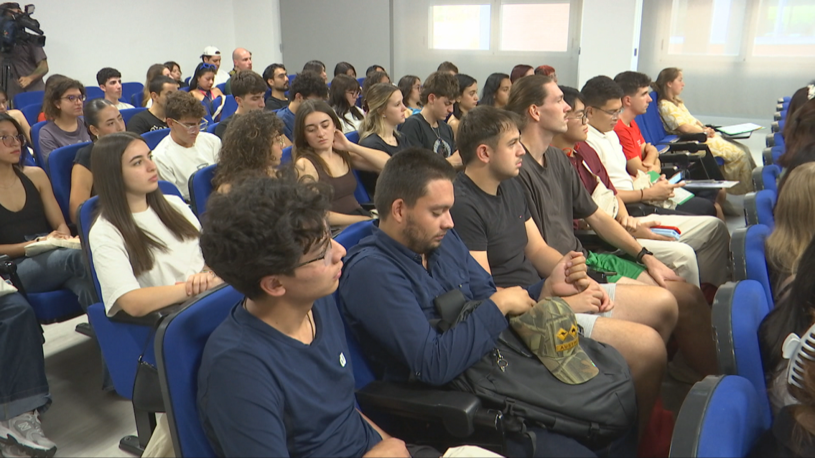 Numerosos jóvenes adultos, sentados en sillas azules en un aula moderna, atienden a un evento mientras una cámara profesional graba la escena. Luz natural ilumina la sala.