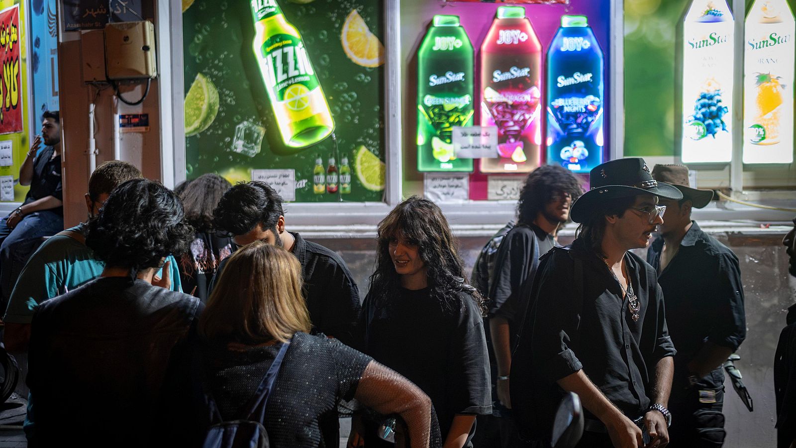 Jóvenes iraníes reunidos en una calle céntrica de Teherán la noche del 11 de septiembre de 2026, rodeados de luminosos anuncios de bebidas.