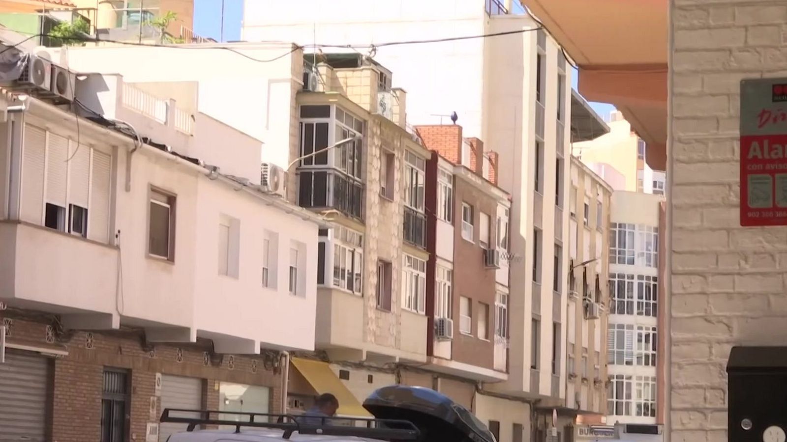 Calle urbana soleada con edificios, furgoneta blanca y cartel de "Alarma Securitas Direct" en pared de ladrillo blanco.