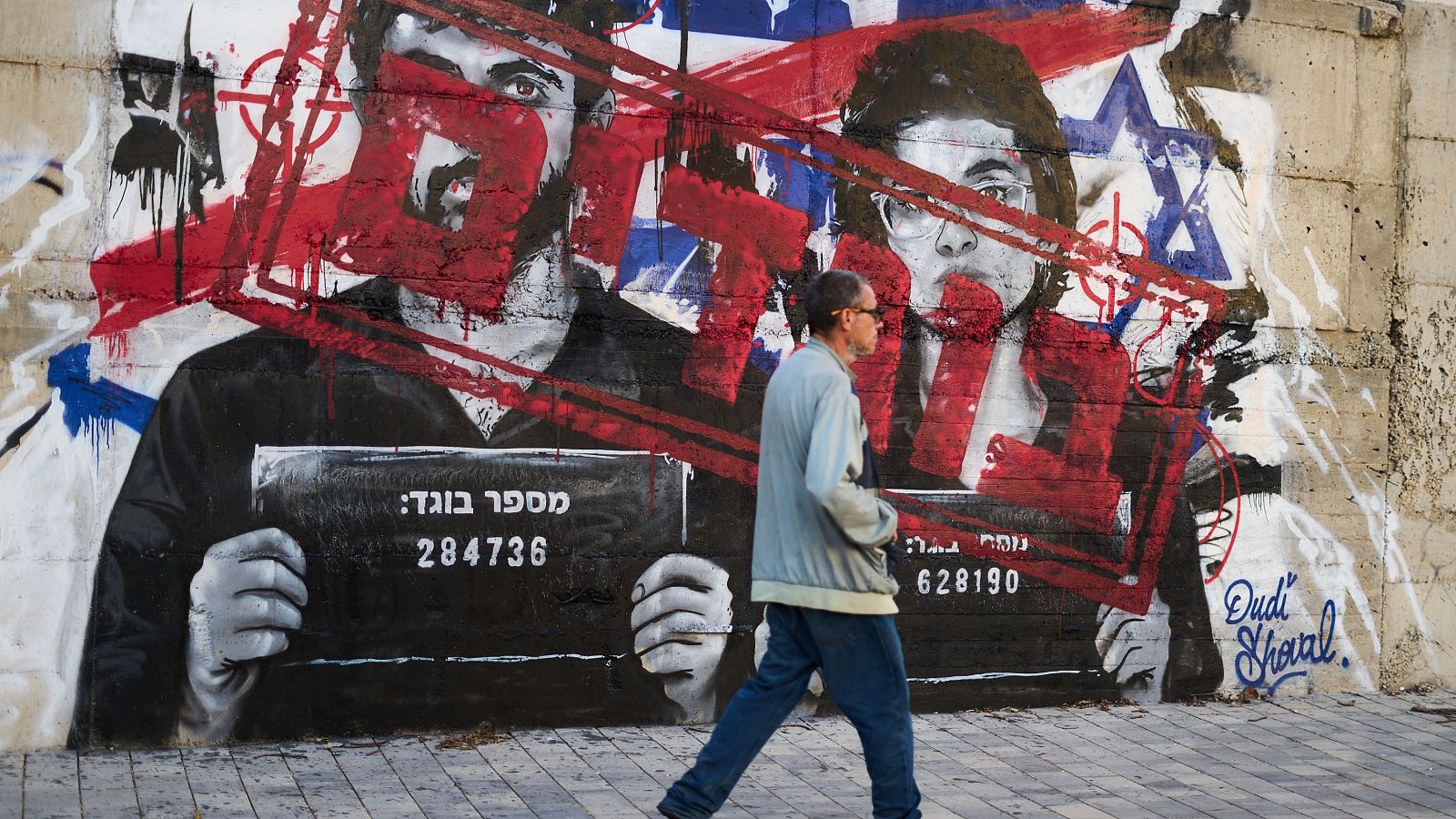 Grafiti en hormigón con retratos de Yuval Abraham y Rachel Szor, marcados con "X" rojas y símbolos de la bandera de Israel. Sostienen carteles con "מספר בוגד:" y números. Firma "Dudi Shoval" en azul.