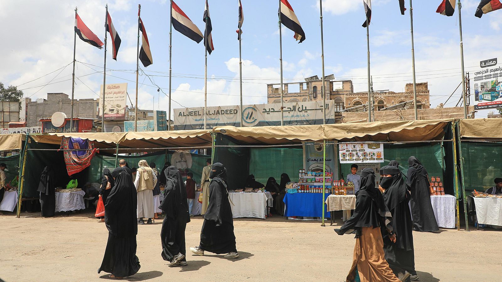 Mercado al aire libre en Yemen con puestos, banderas y edificios tradicionales. Carteles en árabe anuncian "ALJEEL ALJADEED" y "Ceremonias de Boda". Personas vestidas con abayas y niqabs transitan entre la mercancía.
