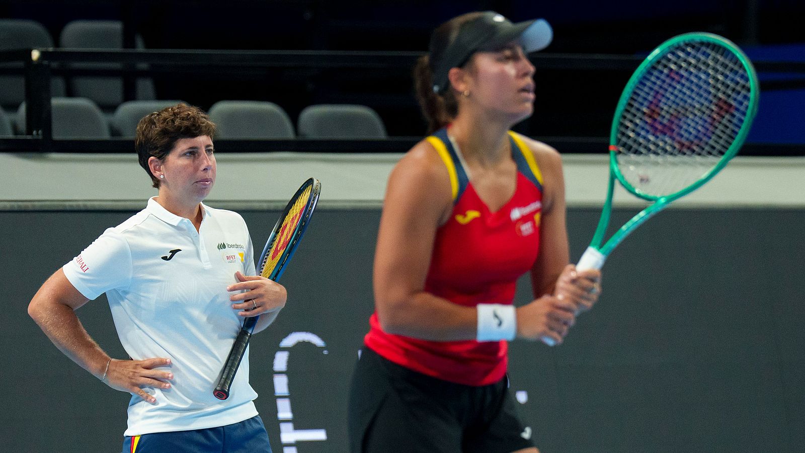 Jessica Bouzas y Carla Suárez, tenistas españolas, entrenan en una cancha cubierta. Ambas portan raquetas y vestimenta deportiva con logos.