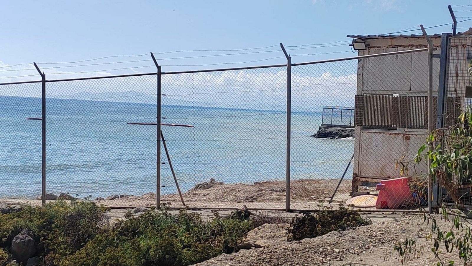 Paisaje costero con valla de seguridad, mar azul verdoso, espigón de hormigón y edificación modular con foco de luz. Terreno árido y rocoso en primer plano.