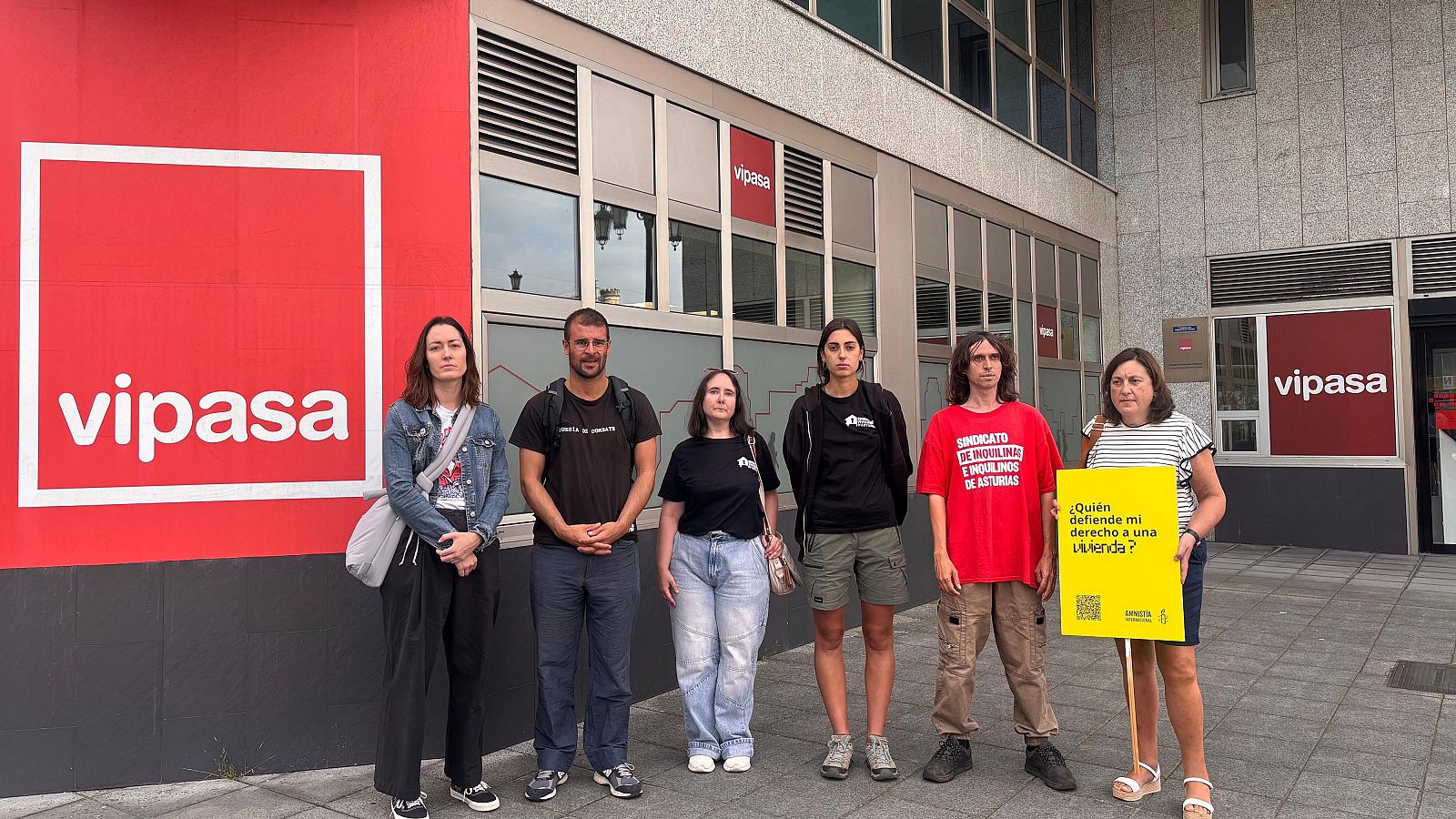 Seis personas frente a un edificio con logo "vipasa". Una mujer porta un cartel amarillo de Amnistía Internacional sobre el derecho a la vivienda.