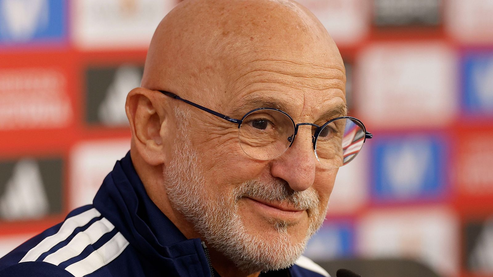 Pierluigi Collina, calvo, con barba canosa y gafas redondas, sonríe. Lleva una chaqueta deportiva azul con tres franjas blancas de Adidas. Fondo de panel de prensa.