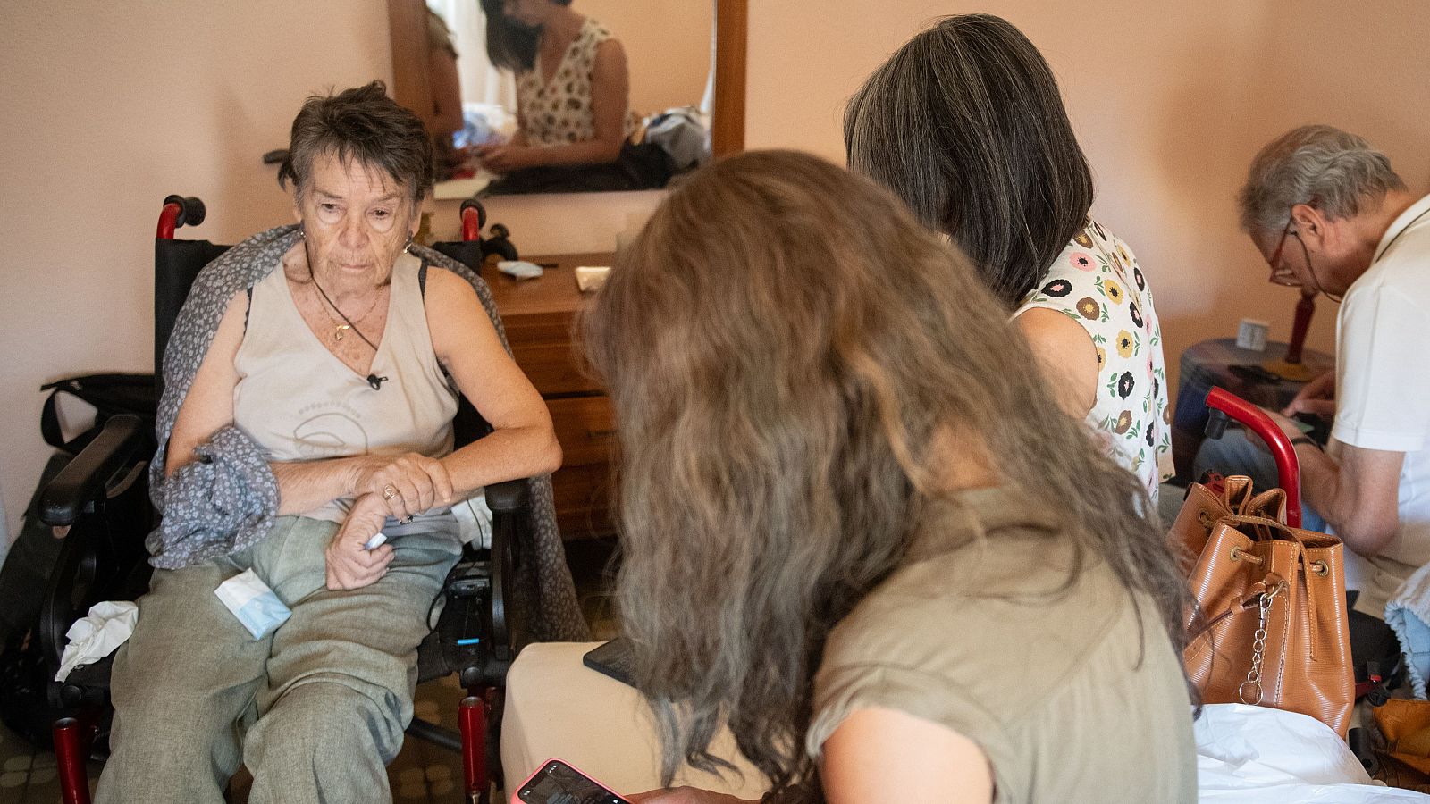 Maricarmen, sentada en silla de ruedas, con micrófono, rodeada de personas en un interior. Expresión de preocupación.