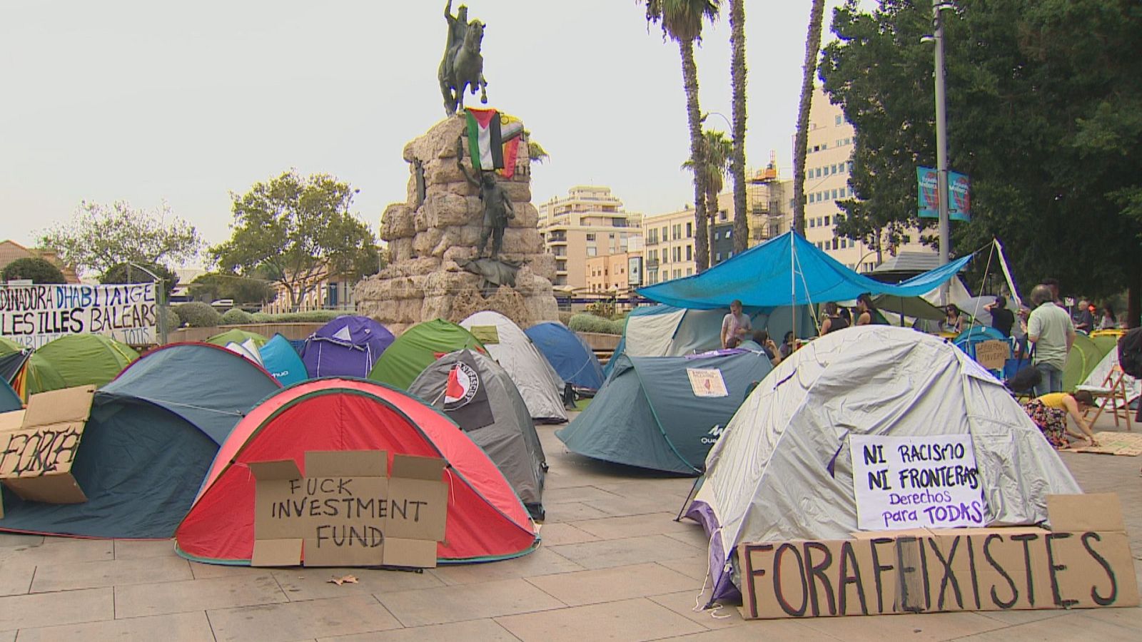 Campamento de protesta en Palma de Mallorca con tiendas de campaña y carteles como 'FUCK INVESTMENT FUND' y 'FORA FEIXISTES' ante el monumento al Rey Jaume I.