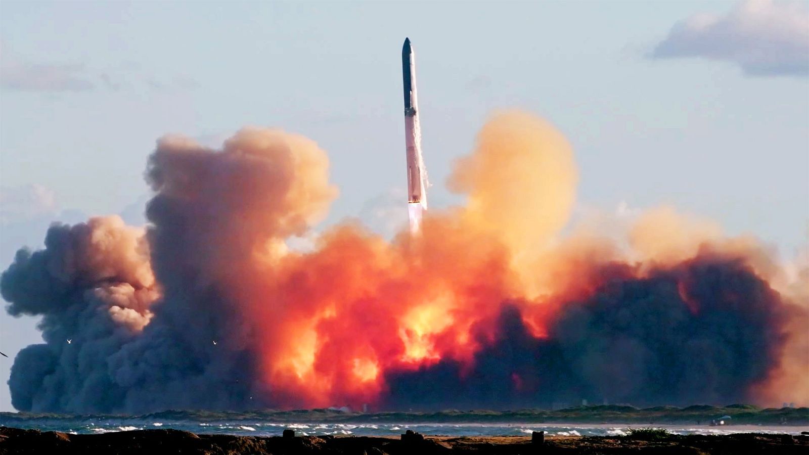 Un gigantesco cohete espacial se eleva dejando una densa nube de humo y fuego sobre la costa en un día despejado.
