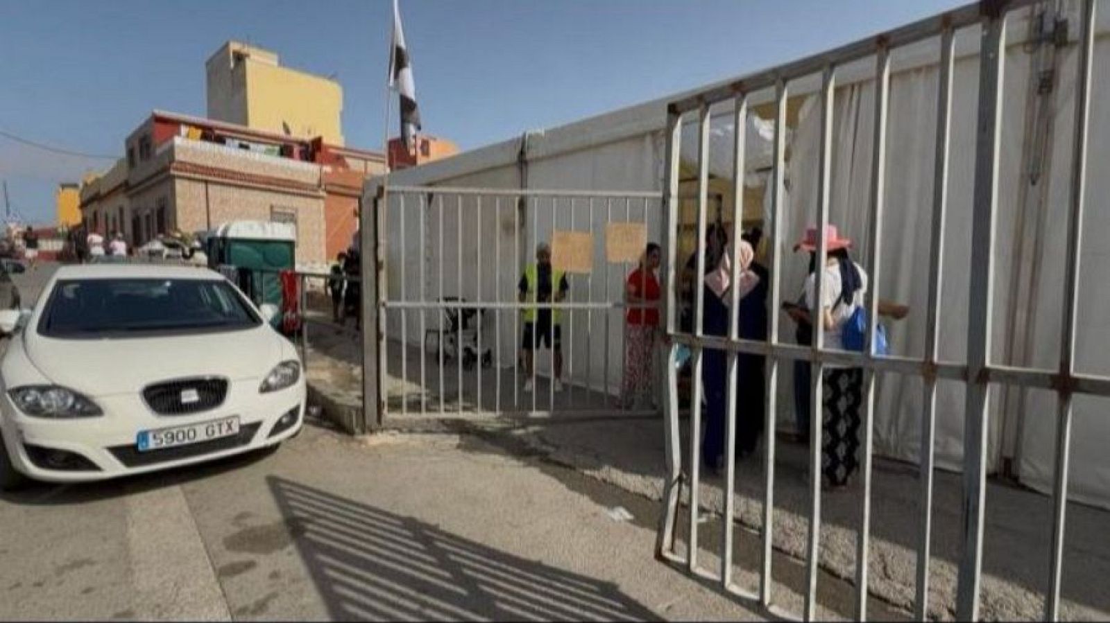 Calle soleada en Ceuta con la entrada a un recinto cercado que cuenta con una carpa blanca y la bandera local, un grupo de personas en el acceso y un coche SEAT León blanco aparcado en la calzada.
