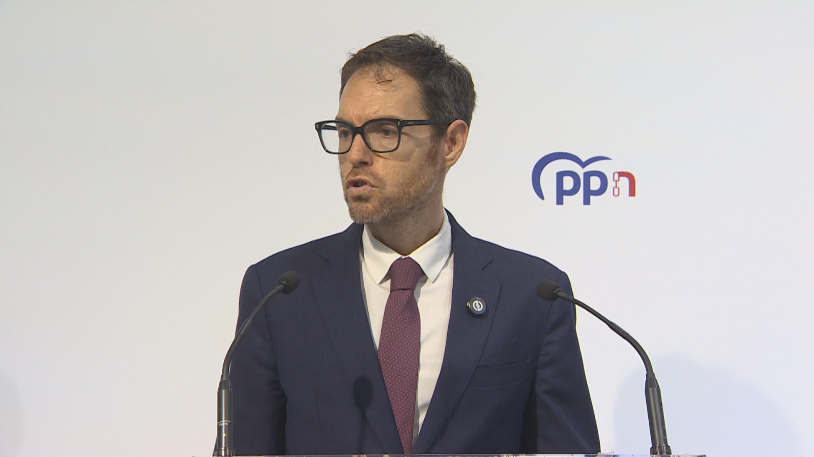 Hombre con traje azul y corbata burdeos comparece en rueda de prensa ante los micrófonos.