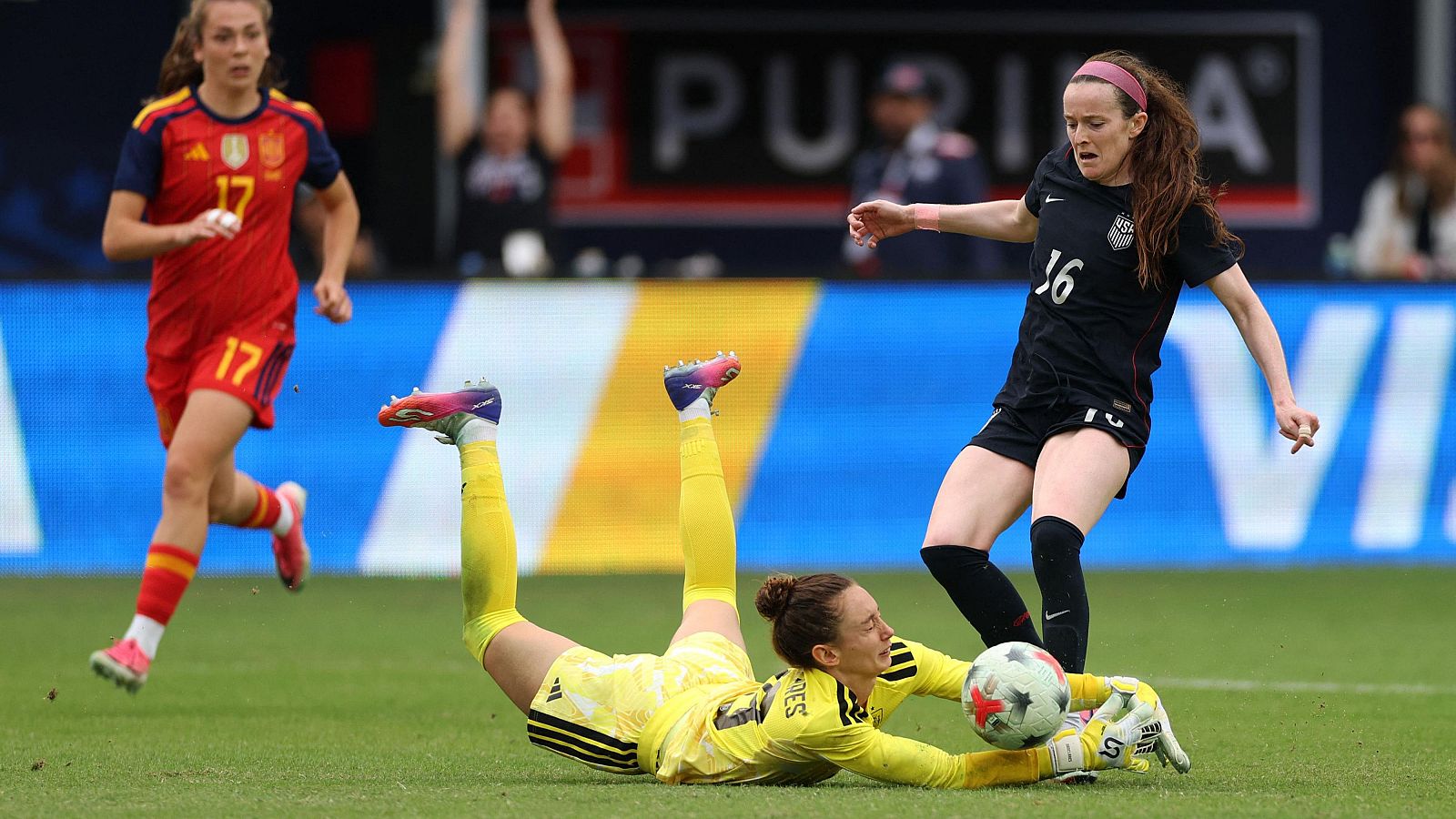 La portera española Misa Rodríguez se desliza en el césped para detener un balón disputado por la estadounidense Rose Lavelle en un partido de fútbol.