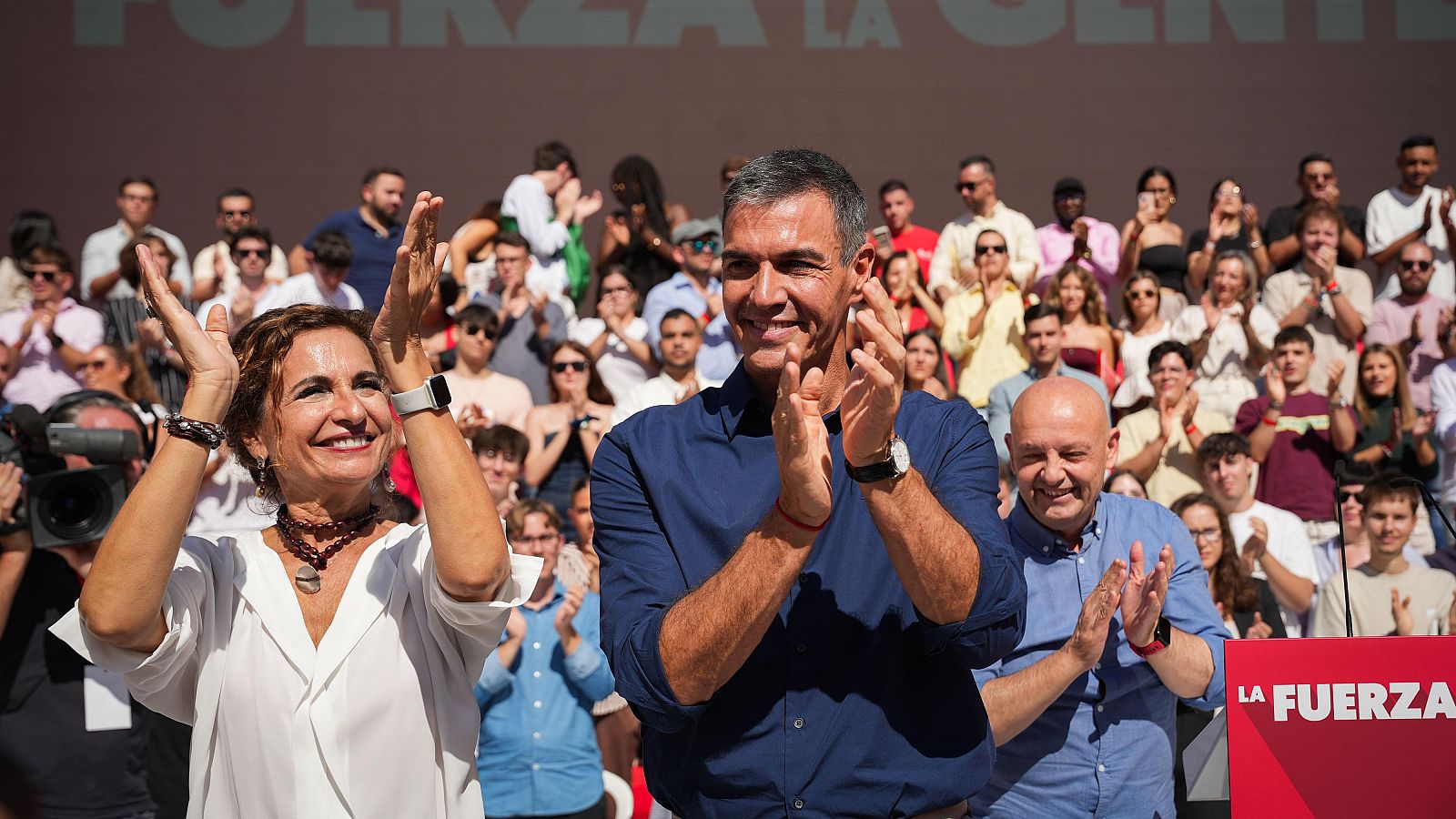 Pedro Sánchez, María Jesús Montero y Francisco Rodríguez García aplauden sonrientes en un acto público al aire libre ante una grada llena de asistentes.