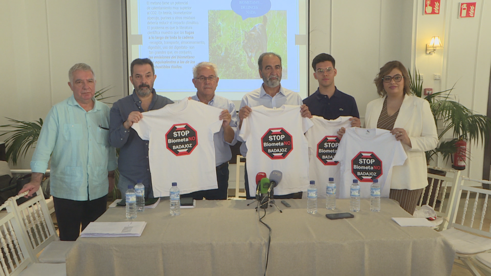 Seis pessoas de pé numa conferência de imprensa mostram camisetas brancas com o lema «STOP BiometaNO BADAJOZ» atrás de uma mesa com microfones e documentos.