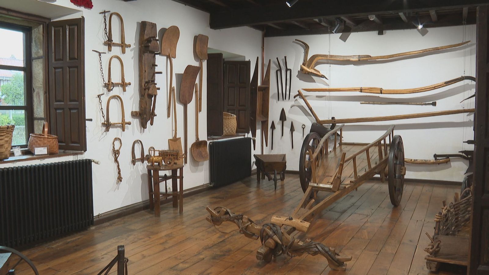 Sala de museo etnográfico con suelo de madera y paredes blancas donde se exhiben un carro tradicional, arados de madera y diversos aperos agrícolas colgantes.