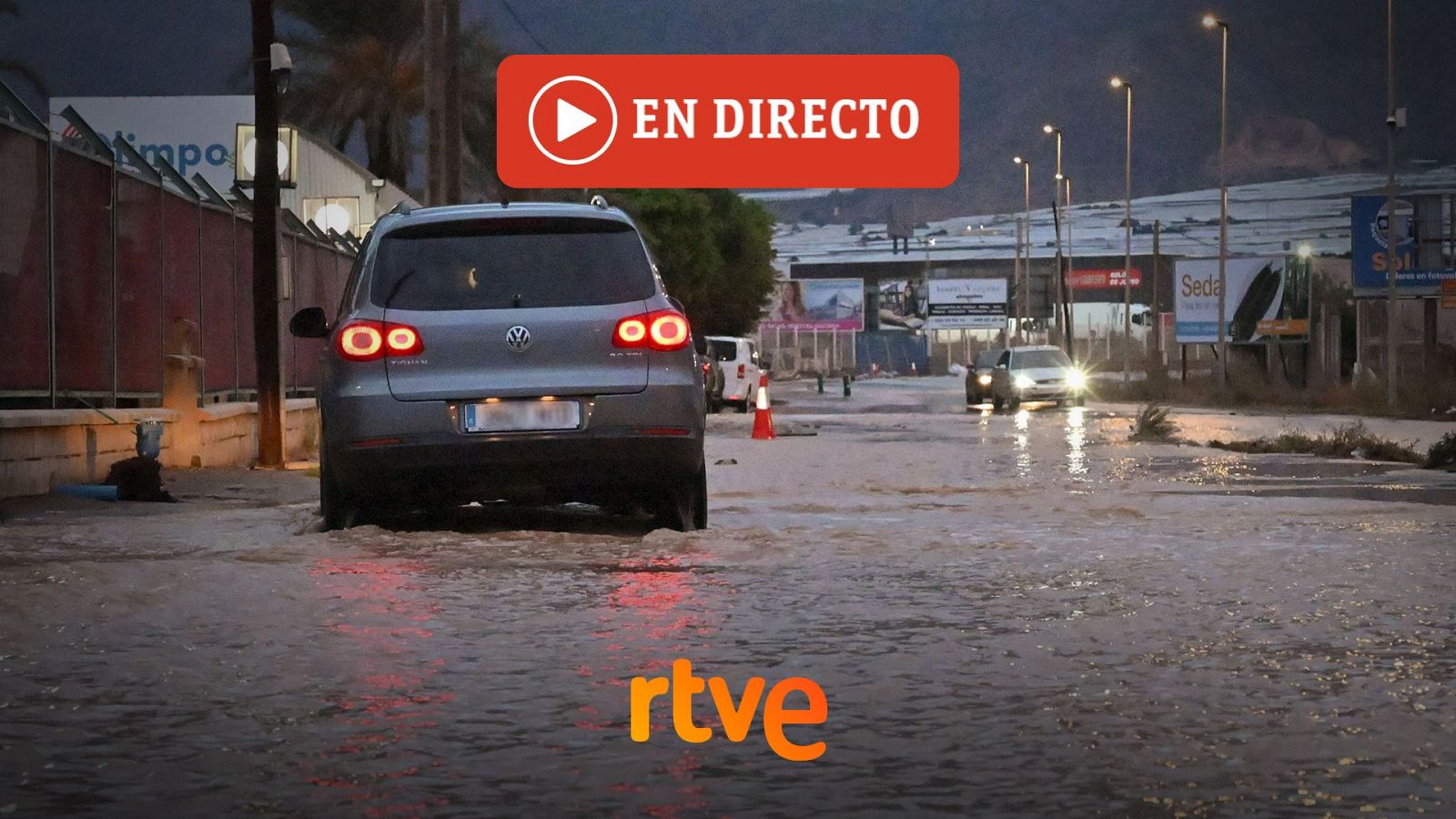 Un todocamino gris circula por una calle industrial inundada y embarrada, con el agua cubriendo la calzada y las luces traseras encendidas.