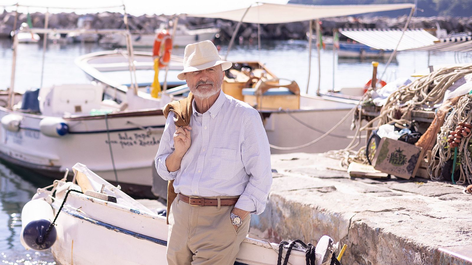 Arturo Pérez-Reverte de pie en un muelle pesquero con camisa clara, pantalones beige, sombrero y chaqueta al hombro.