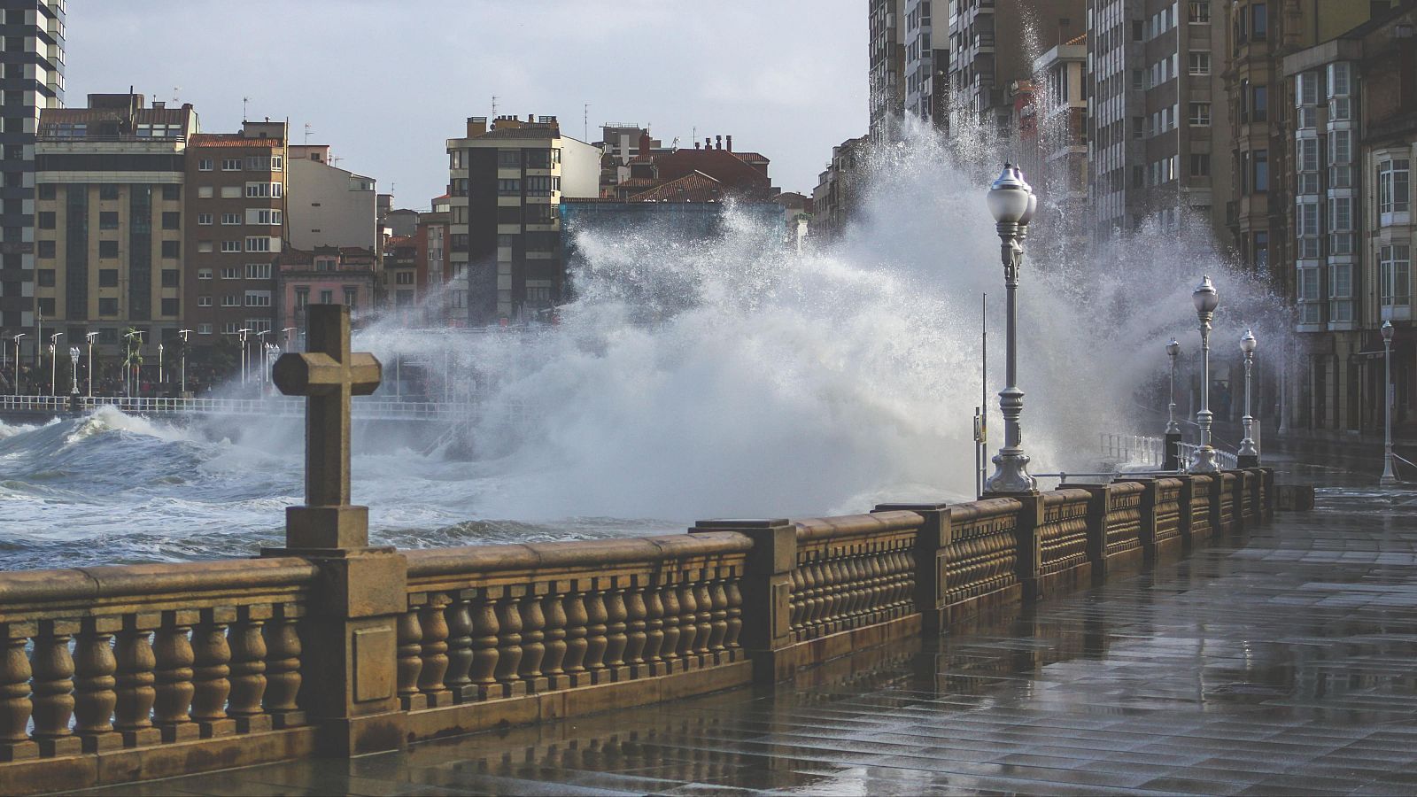 Olas rompiendo con fuerza en el paseo marítimo de Gijón bajo un cielo nublado debido al viento del noroeste.