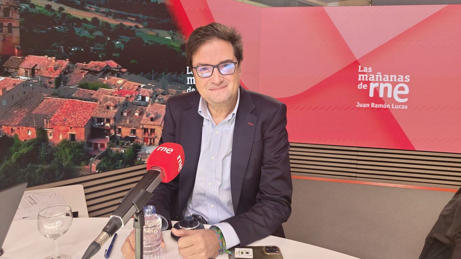 El periodista Íñigo Alfonso sonríe ante un micrófono de radio en un estudio con paneles decorativos y el logotipo del programa de RNE.
