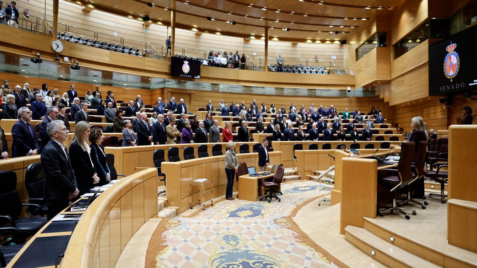 Pleno en el hemiciclo del Senado de España con personas de pie guardando silencio junto a la mesa presidencial y una pantalla con la palabra 'SENADO'.