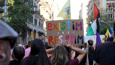 Manifestación urbana con cartel "existIM RESISTIM" y corazones. Se ven la Estelada groga, bandera de Palestina y bandera morada. Calle con edificios, árboles y multitud. Manifestación urbana con cartel "existIM RESISTIM" y corazones. Se ven la Estelada groga, bandera de Palestina y bandera morada. Calle con edificios, árboles y multitud.