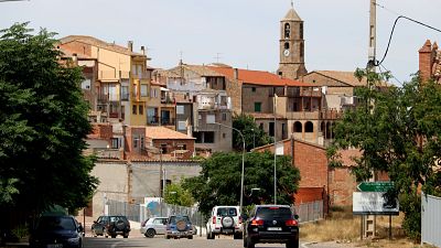 Vista de la entrada a Bellaguarda con carretera, vehículos, edificios tradicionales y un campanario de piedra con reloj al fondo. Vista de la entrada a Bellaguarda con carretera, vehículos, edificios tradicionales y un campanario de piedra con reloj al fondo.