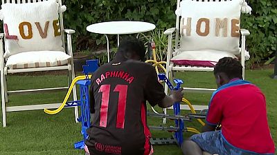 Niños jugando con un circuito de coches en un jardín con mobiliario blanco.  Uno viste camiseta con el nombre "RAPHINHA" y el número 11.