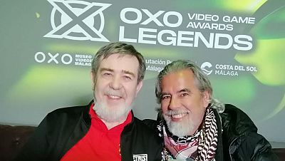 Dos hombres sonríen juntos frente a un panel con logos. Uno de ellos, con camisa roja y chaqueta con el logo de Tetris, posa junto a otro con chaqueta negra y bufanda estampada. Dos hombres sonríen juntos frente a un panel con logos. Uno de ellos, con camisa roja y chaqueta con el logo de Tetris, posa junto a otro con chaqueta negra y bufanda estampada.