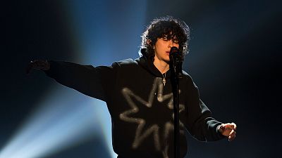 Cantante con sudadera negra y capucha actuando en un escenario iluminado. Pelo rizado oscuro y micrófono.