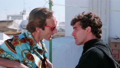 Escena de película con Eusebio Poncela, camisa estampada y gafas rojas, mirando a Antonio Banderas, quien viste ropa oscura. Fondo con tendedero. Escena de película con Eusebio Poncela, camisa estampada y gafas rojas, mirando a Antonio Banderas, quien viste ropa oscura. Fondo con tendedero.