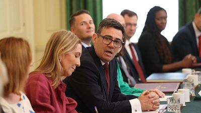 Andy Burnham, con gafas y traje, lidera una reunión formal. Participan otras personas, algunas observando atentamente. La mesa tiene documentos y vasos. Andy Burnham, con gafas y traje, lidera una reunión formal. Participan otras personas, algunas observando atentamente. La mesa tiene documentos y vasos.