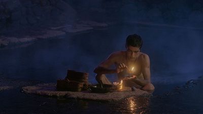 Un hombre joven de piel morena, con el torso desnudo, realiza un ritual en un entorno acuático oscuro, sosteniendo una llama y con un cuenco metálico frente a él. La luz del fuego ilumina la escena. Un hombre joven de piel morena, con el torso desnudo, realiza un ritual en un entorno acuático oscuro, sosteniendo una llama y con un cuenco metálico frente a él. La luz del fuego ilumina la escena.