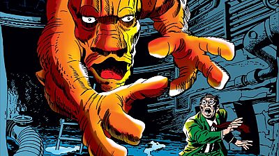 Monstruo naranja gigante amenaza a un hombre en traje verde y corbata roja en una escena oscura y claustrofóbica de un cómic de terror.