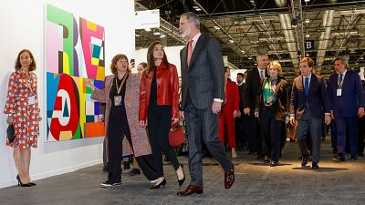 Los Reyes de España recorren la inauguración de ARCO en IFEMA. Él lleva traje gris y corbata rosa; ella, chaqueta roja y pantalones negros. Se observa una obra con letras grandes y coloridas.