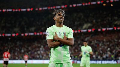 Celebración de gol de un jugador de fútbol con camiseta verde claro y pantalón a juego, brazos cruzados y expresión alegre. Estadio lleno y líneas del campo visibles. Celebración de gol de un jugador de fútbol con camiseta verde claro y pantalón a juego, brazos cruzados y expresión alegre. Estadio lleno y líneas del campo visibles.