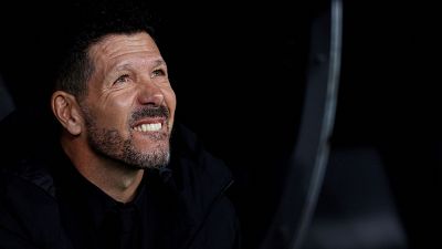 Diego Simeone, con chaqueta negra, muestra una leve sonrisa que podría interpretarse como irónica. Mira hacia arriba y a la derecha sobre un fondo oscuro.
