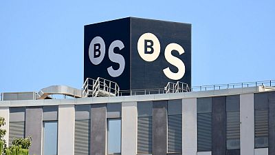 Logotipo "BS" blanco sobre fondo negro en la azotea de un edificio moderno con fachada gris y blanca. Se observan ventanas y estructuras metálicas.