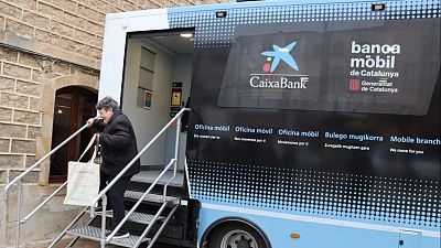 Mujer mayor subiendo escaleras a oficina bancaria móvil de CaixaBank. Vehículo con logotipos de CaixaBank y Generalitat de Catalunya. Mujer mayor subiendo escaleras a oficina bancaria móvil de CaixaBank. Vehículo con logotipos de CaixaBank y Generalitat de Catalunya.
