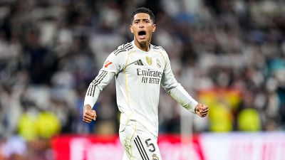 Un jugador de fútbol, con la camiseta blanca de su equipo y el número 5, celebra un gol con los brazos en alto, ante un fondo difuminado con público.