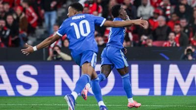 Dos jugadores de fútbol con uniformes azules y detalles blancos celebran un gol, uno de espaldas con el dorsal 10 y el nombre "MBAPPE", y el otro de frente con los brazos extendidos, ambos con zapatillas rosas.
